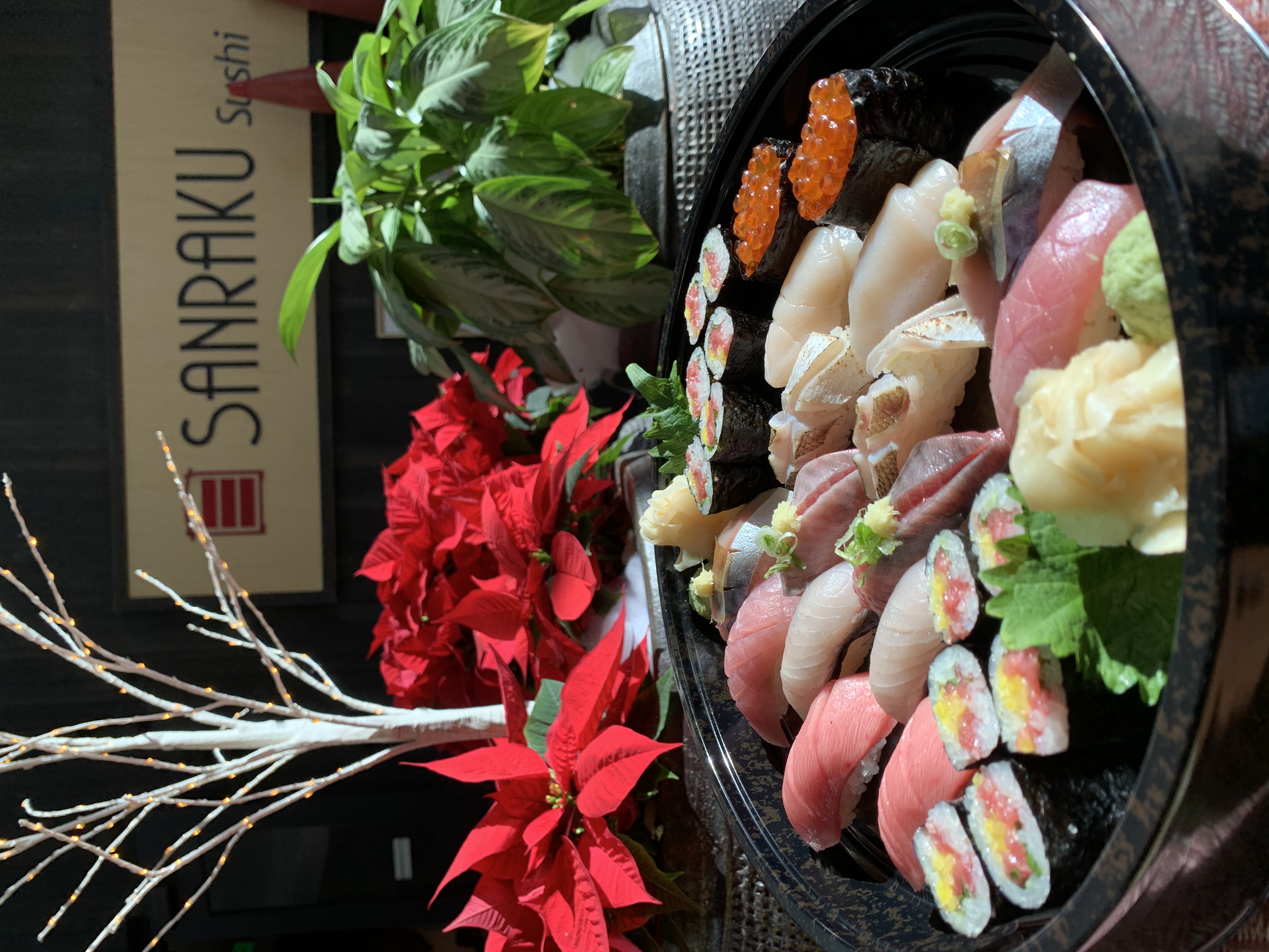 A beautifully arranged platter of sushi featuring various types, including nigiri and maki, presented in front of a festive display with red poinsettias and a decorative tree at Sanraku Sushi restaurant.